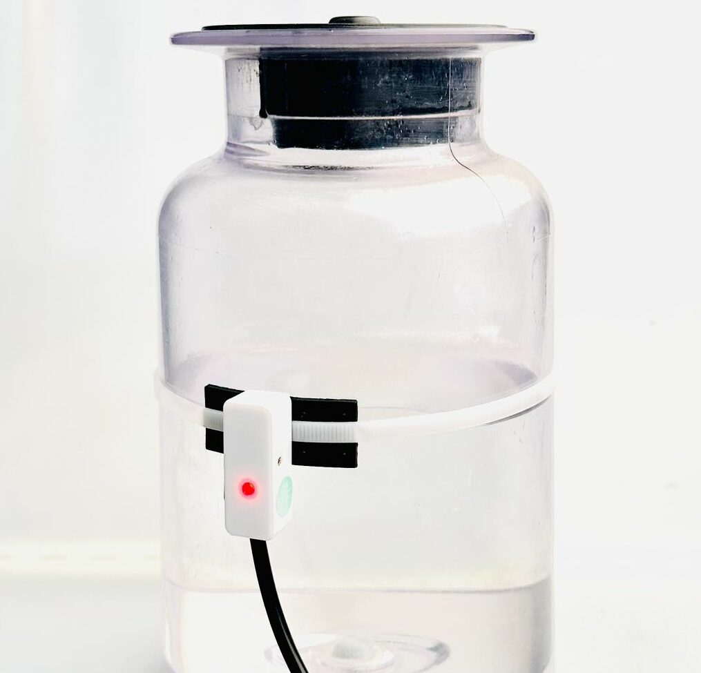 A lit up non contact fluid level sensor strapped to a jar, determining that the fluid level within has reached the required height.